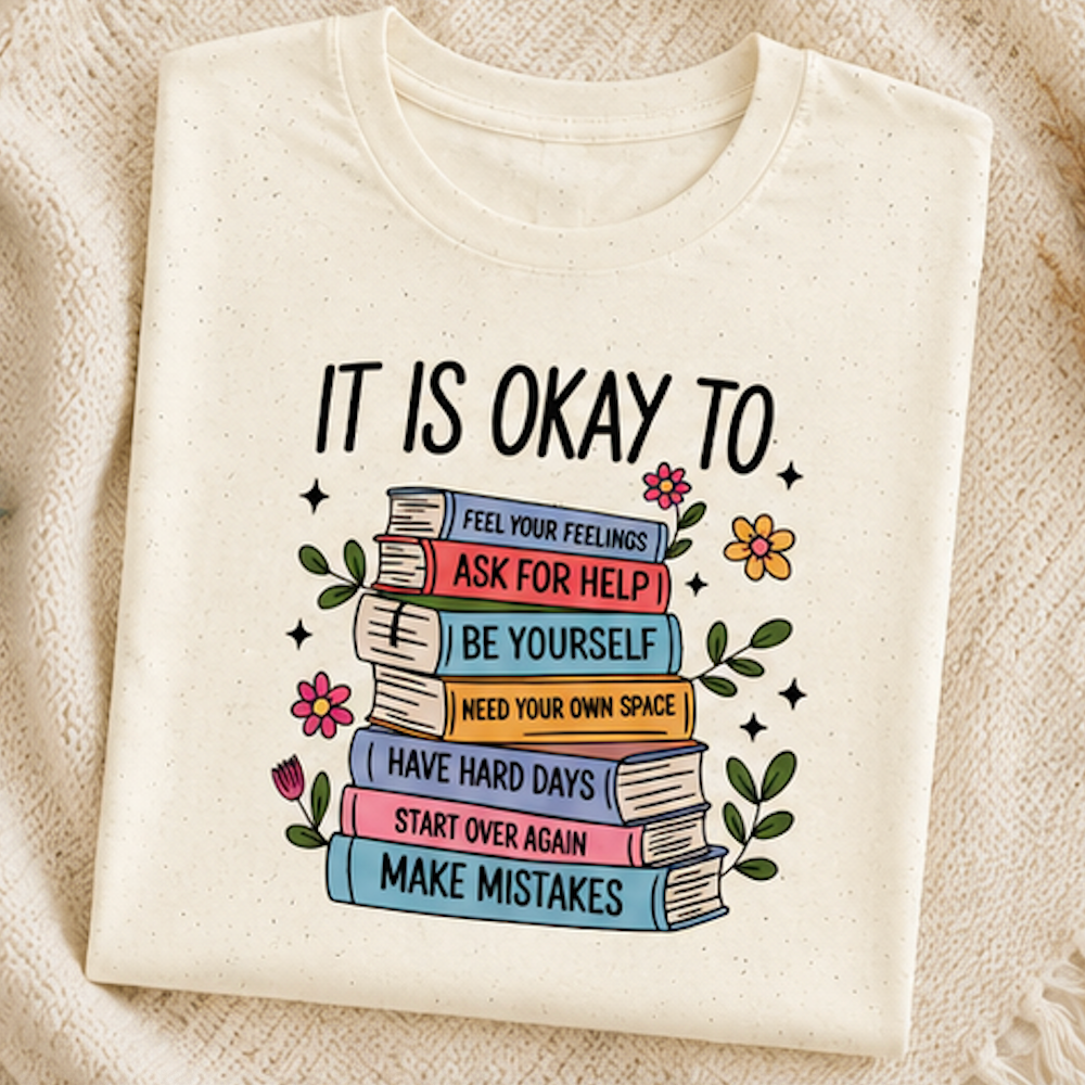 An off-white crewneck t-shirt laid flat on a textured beige knit blanket. The shirt features a graphic print of a stack of colorful books with motivational self-care phrases like 'ASK FOR HELP', 'BE YOURSELF', and 'MAKE MISTAKES'.