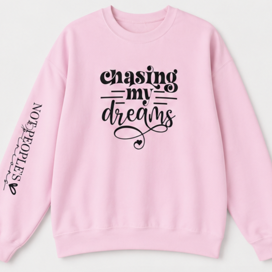 A light pink crewneck sweatshirt with black graphic text on the front reading 'chasing my dreams' and 'NOT PEOPLES' printed down the left sleeve, shown against a light gray background.