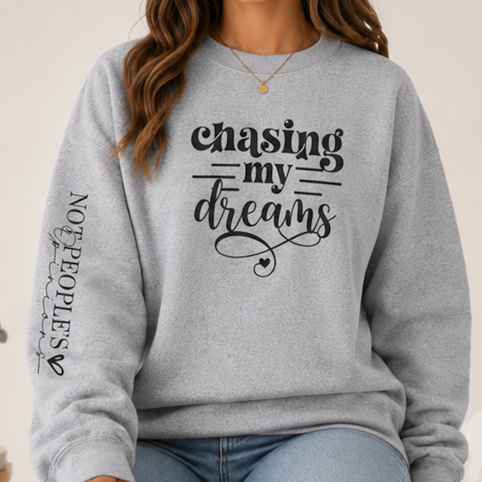 A woman wearing a light heather gray crewneck sweatshirt with black graphic text reading 'Chasing my dreams' on the front and 'NOT PEOPLES' with a heart on the sleeve, paired with light blue jeans.