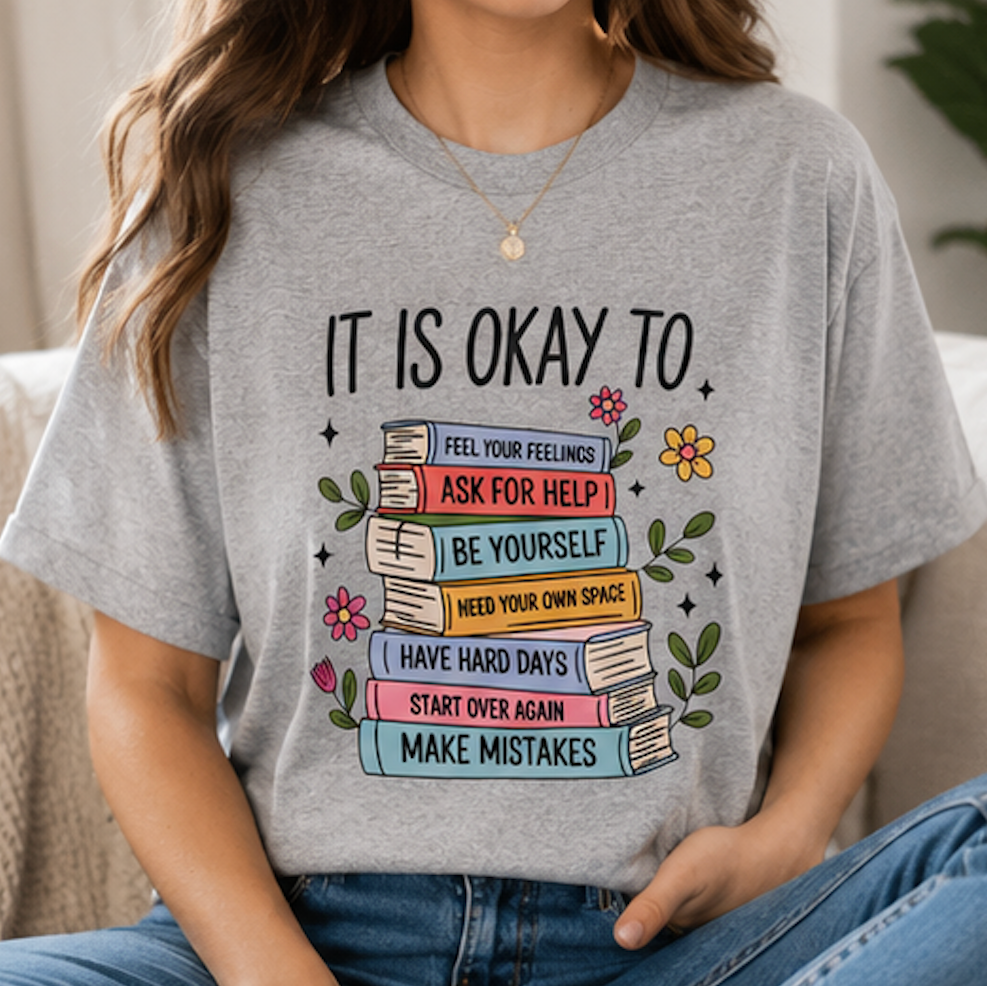A woman wearing a heather gray graphic T-shirt with a colorful book stack design and the text 'IT IS OKAY TO.