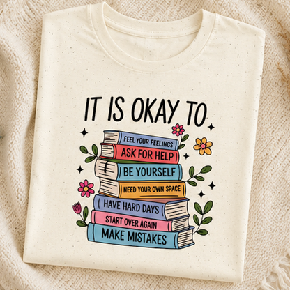 An off-white crewneck t-shirt laid flat on a textured beige knit blanket. The shirt features a graphic print of a stack of colorful books with motivational self-care phrases like 'ASK FOR HELP', 'BE YOURSELF', and 'MAKE MISTAKES'.