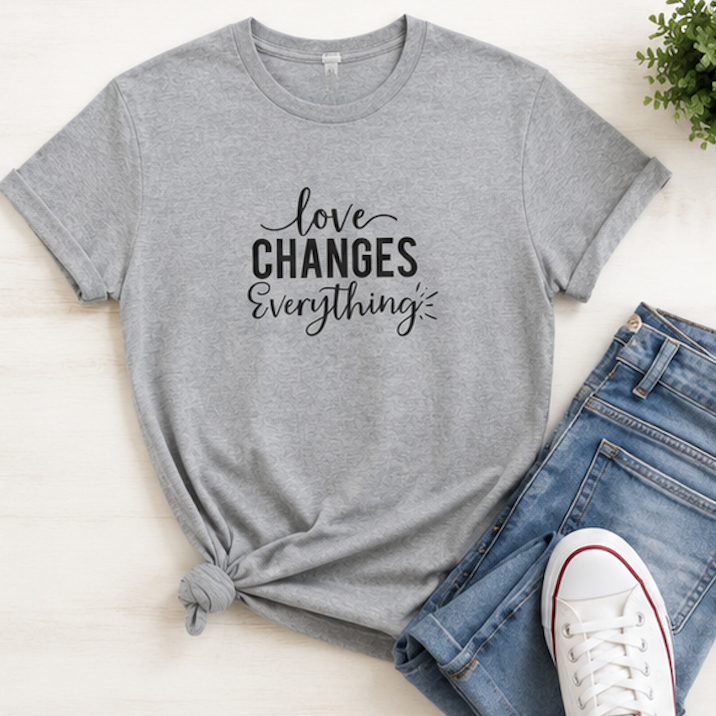 A heather gray short-sleeve graphic T-shirt laid flat on a light wood surface, styled with blue jeans, a white sneaker, and a small plant. The shirt has black text reading 'love CHANGES everything' across the chest.
