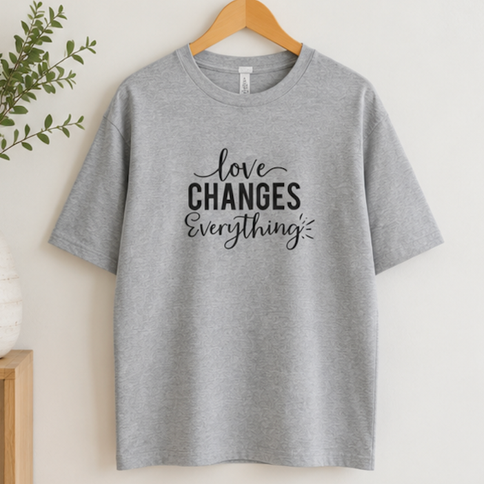 A heather gray short-sleeve t-shirt hanging on a wooden hanger against a light beige wall, featuring the black printed text 'love CHANGES Everything' on the front.