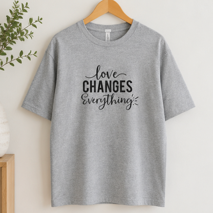 A heather gray short-sleeve t-shirt hanging on a wooden hanger against a light beige wall, featuring the black printed text 'love CHANGES Everything' on the front.