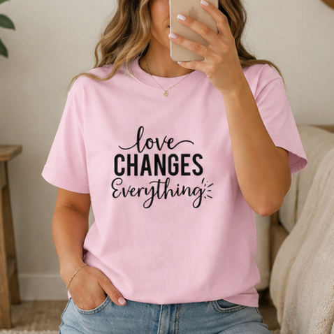 A woman takes a mirror selfie wearing a light pink crewneck t-shirt with the black printed text 'love CHANGES Everything'.