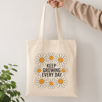 A cream canvas tote bag with a daisy graphic and the text 'KEEP GROWING EVERY DAY' in black, held by a person's hand against a light gray wall. A green plant is visible in the lower-left corner.