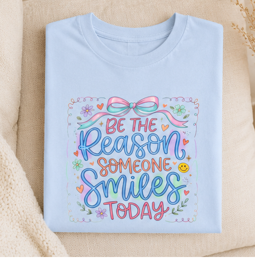 A four-panel collage showing a light blue graphic T-shirt in different styling contexts: worn by a model, laid flat, and worn again in a seated setting. The shirt has a colorful decorative bow and floral design with the phrase “Be the Reason Someone Smiles Today.