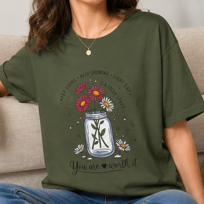 A four-panel collage showing an olive green graphic T-shirt worn by a model in different settings, plus a flat lay of the same shirt. The shirt features a mason jar design with flowers, a cross, and the text 'You are worth it'.