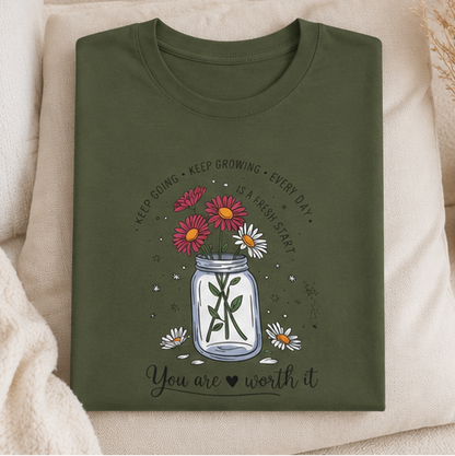 A four-panel collage showing an olive green graphic T-shirt worn by a model in different settings, plus a flat lay of the same shirt. The shirt features a mason jar design with flowers, a cross, and the text 'You are worth it'.