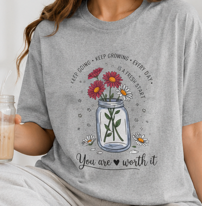 A four-panel collage showing a heather gray graphic T-shirt worn by a person in two lifestyle settings and laid flat on a couch. The shirt has a floral vase design with red flowers, a blue monogram 'JK', and the text 'You are worth it'.